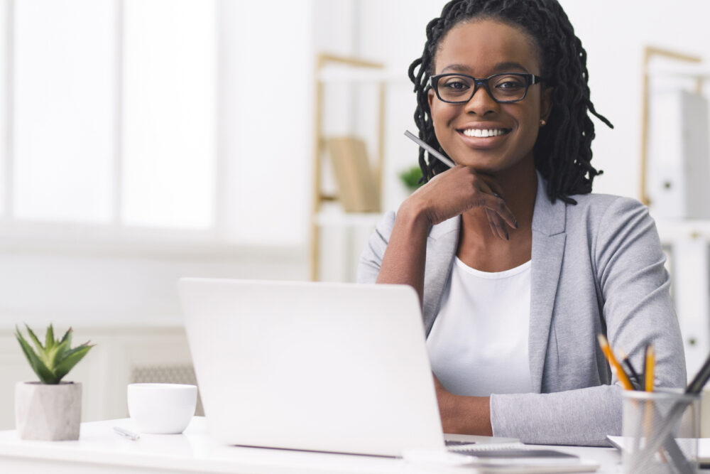 Female Entrepreneurship. Afro Businesswoman Smiling At Camera Touching Chin At Workplace. Empty Space